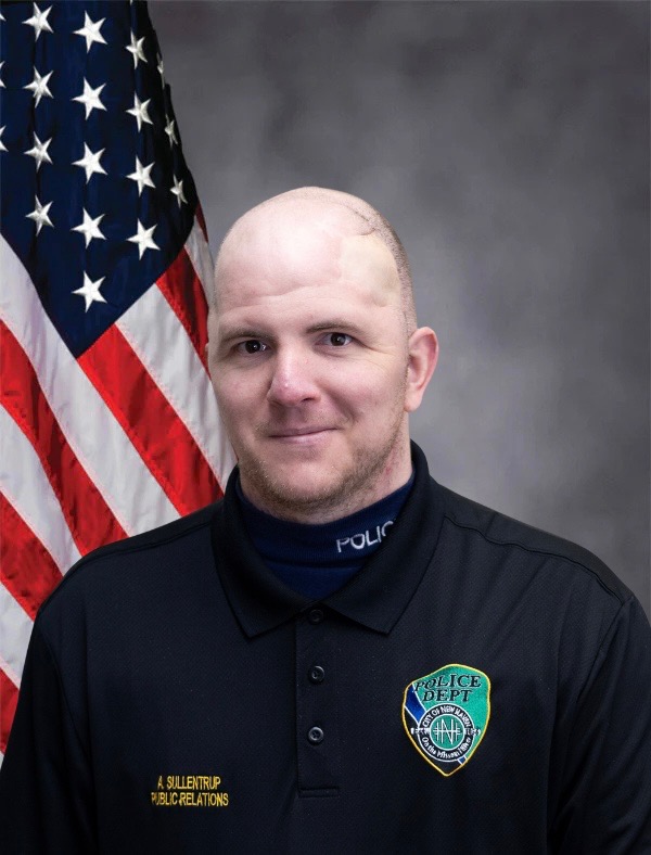Bald man in a dark police uniform with a department badge, standing in front of a U.S. flag backdrop.